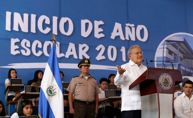 Ein Mann steht an einem Pult mit Mikrofonen und einer Flagge und spricht zu einer Menge an einer Schule, mit einem Banner mit Text und einem Bild eines Gebäudes im Hintergrund.