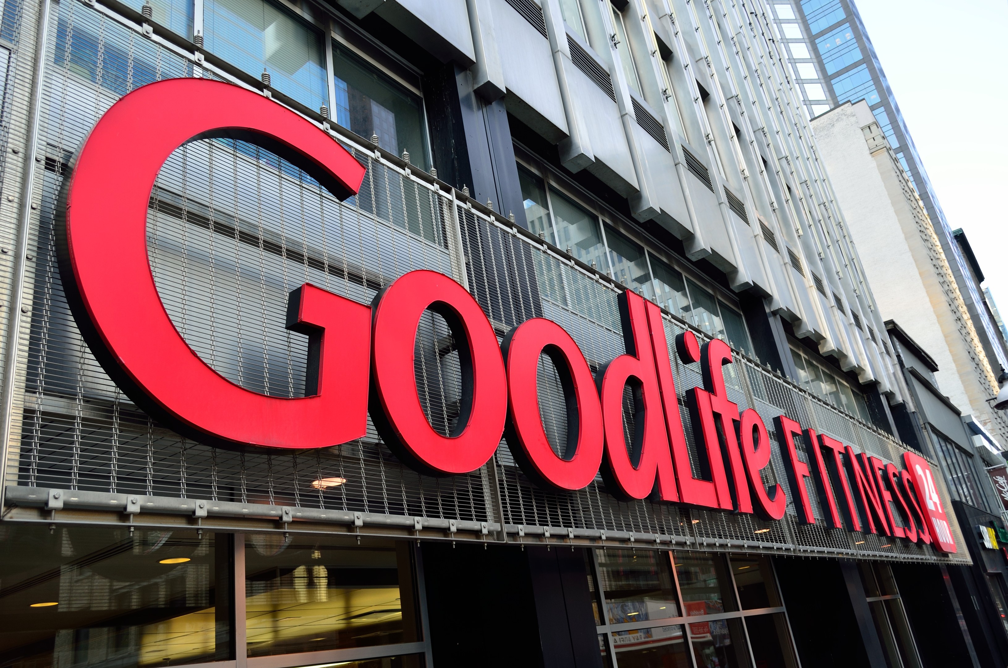 Außenansicht eines Goodlife Fitness Studios in New York City mit Glaswänden, einem Namensschild, benachbarten Gebäuden auf der rechten Seite und Himmel im Hintergrund.