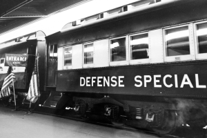 Ein Schwarz-Weiß-Foto eines Zuges mit der Aufschrift 'Defense Special', der auf den Schienen steht, mit zwei Flaggen auf der linken Seite und einer Decke darüber.