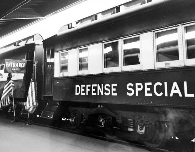 Ein Schwarz-Weiß-Foto eines Zuges mit der Aufschrift 'Defense Special', der auf den Schienen steht, mit zwei Flaggen auf der linken Seite und einer Decke darüber.