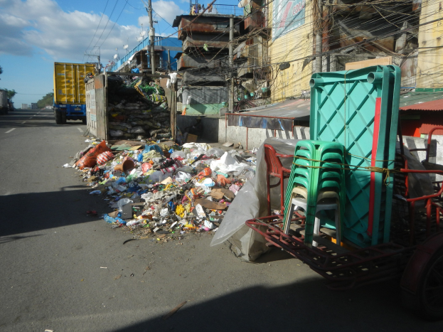Ein Lastwagen neben einem Müllhaufen auf einer Straße geparkt, mit einem Wagen rechts daneben, der mit Plastikstühlen beladen ist, und im Hintergrund Gebäude, Strommasten, Bäume und ein bewölkter Himmel.