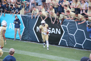 Ein Fußballspieler läuft mit dem Ball auf einem grünen Feld vor einer Menge, einige halten Kameras und tragen Helme, mit einem Banner im Hintergrund.