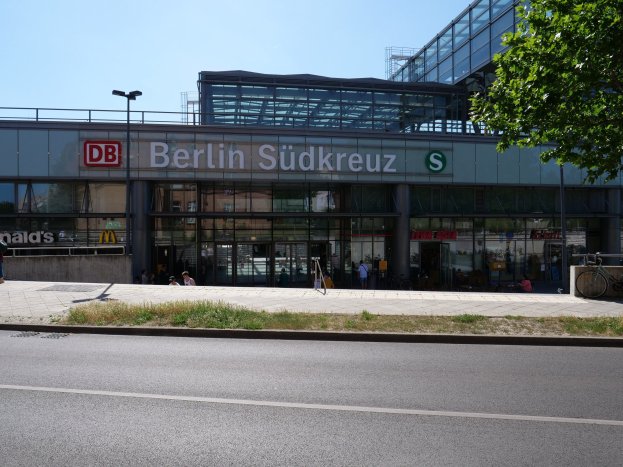 Ein großes Glaswand-Bahnhofgebäude mit Text darauf, umgeben von Straßenlaternen, Lampen, Fahrzeugen, Fußgängern, Fahrrädern, Bäumen und einem klaren blauen Himmel.