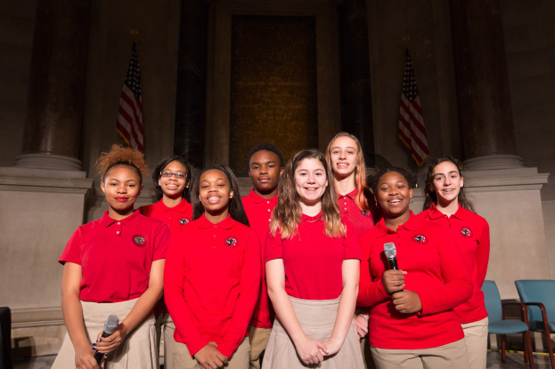 Gruppe junger Frauen in roten Shirts, die strahlend Mikrofone halten und vor Stühlen, Säulen, Fahnen und einer Wand stehen, wahrscheinlich Teil eines nationalen Jugendchors.