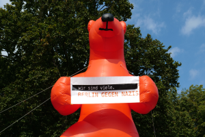 Ein aufblasbarer Bär hält ein Schild mit der Aufschrift "Berlin gegen Nazis" in den Vordergrund, mit Bäumen und Wolken im Hintergrund.