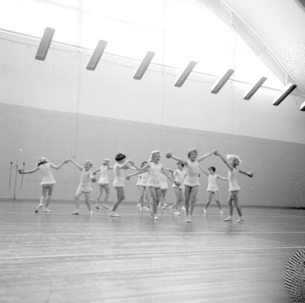 Schwarzes und weißes Bild von jungen Mädchen, die sich an den Händen halten und in einem Studio tanzen, mit einem Ball auf der rechten Seite und Lampen an der Decke, einem Objekt an der Wand im Hintergrund.