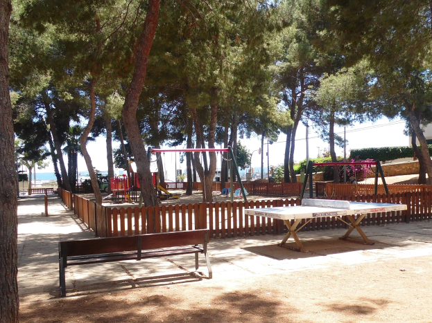 Ein Park mit einem zentralen Tischtennis-Tisch, umgeben von Bänken, einem Zaun, Schaukeln, Pfosten, Pflanzen und Bäumen, vor einem Gewässer und einem klaren blauen Himmel.