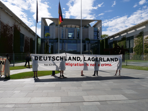 Gruppe von Menschen mit einem Banner 'Deutschland, Lagerland Migration ist kein Verbrechen' vor einem Gebäude mit Fenstern, umgeben von Gras, einem Metallzaun, Pflanzen, Bäumen und Fahnenstangen.