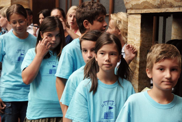 Eine Gruppe von Kindern steht vor einer Wand oder Säule, mit weiteren Personen dahinter und zusätzlichen Säulen und Fenstern im Hintergrund.