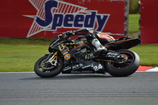 Eine Person in Helm und Schutzkleidung fährt ein Motorrad auf einer Rennstrecke, umgeben von Gras, mit einer Texttafel im Hintergrund.