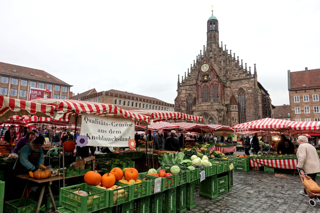 Ein belebter Markt in Nürnberg, Deutschland, mit Ständen voller Obst und Gemüse, Menschen mit Taschen und aufgestellten Zelten, sowie Gebäuden und einem Uhrenturm im Hintergrund unter einem sichtbaren Himmel.