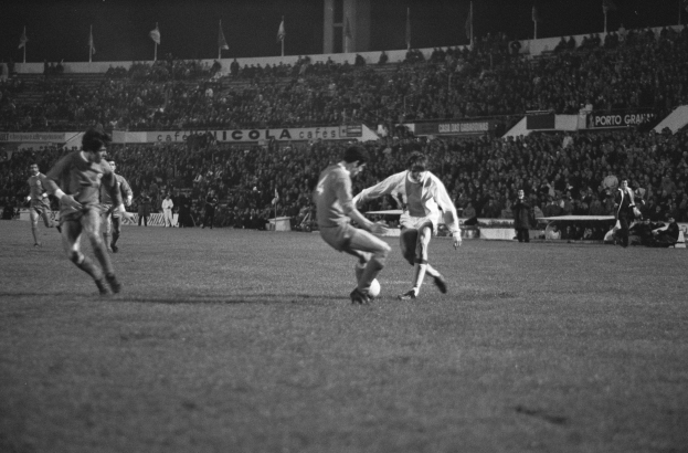 Schwarzes und weißes Bild eines Fußballspiels in einem Stadion mit Spielern auf dem Feld und Zuschauern in den Rängen.