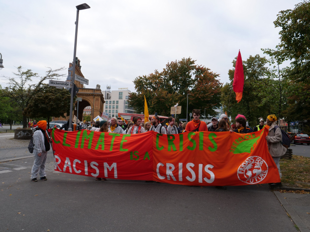 Eine Gruppe von Menschen marschiert eine baumbestandene Straße entlang und hält ein "Klimakrise ist eine Krise"-Schild, mit Fahrzeugen, Gebäuden und einem klaren blauen Himmel im Hintergrund.