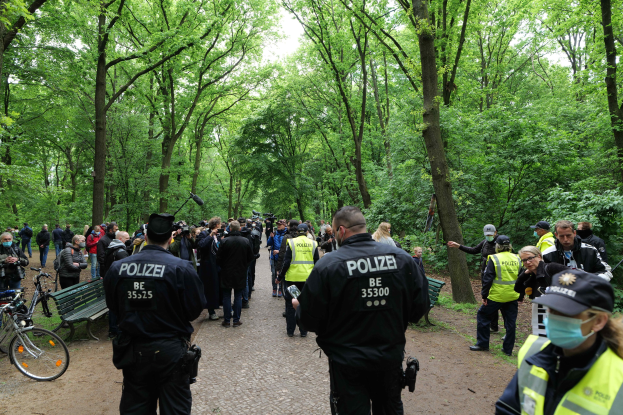 Polizisten vor einer Menge bei einer Outdoor-Veranstaltung, mit Fahrrädern und einer Bank im Vordergrund und Bäumen im Hintergrund.