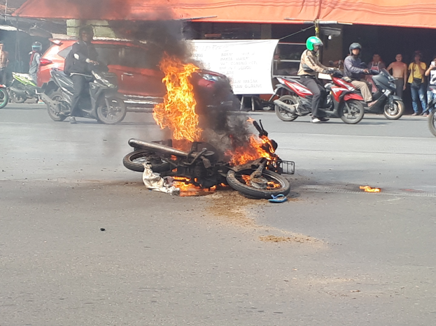 Ein auf der Straße in Flammen stehendes Motorrad mit einem Auto und einem Van im Hintergrund, umgeben von Menschen, einige tragen Helme, eine Fahne mit Text und ein Zelt.