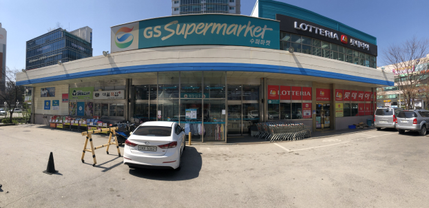 Ein Auto vor einem Geschäft mit einer "GS Supermarkt"-Reklame, umgeben von anderen Fahrzeugen, Verkehrskegeln und Gegenständen, mit Gebäuden, Bäumen und einem klaren blauen Himmel im Hintergrund.