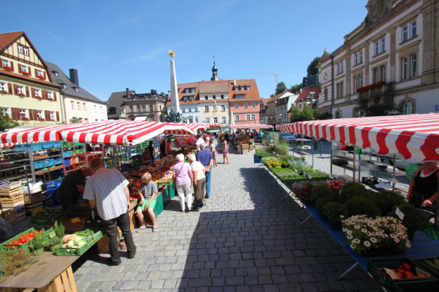 Ein belebter Markt im historischen Zentrum von Heidelberg mit Menschen, die spazieren gehen, auf Bänken sitzen und in der Nähe von Zelten stehen, umgeben von Gemüsekörben, Gebäuden, Bäumen und einem klaren blauen Himmel.