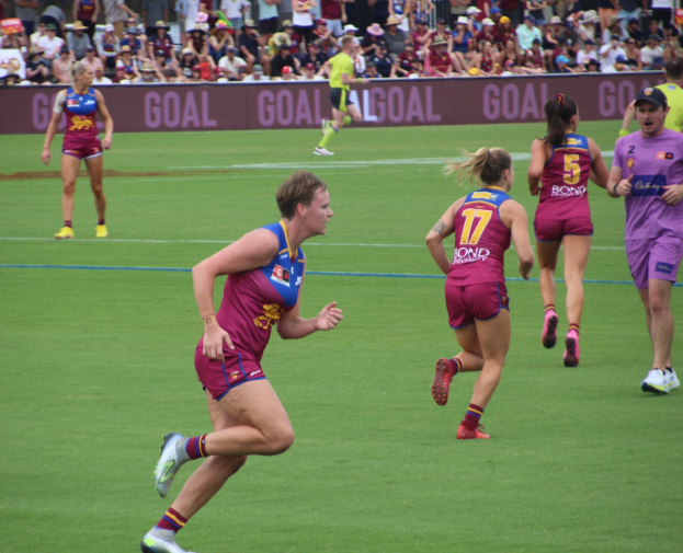 Frauen spielen Australian Rules Football auf einem Rasenfeld mit Zuschauern im Hintergrund.