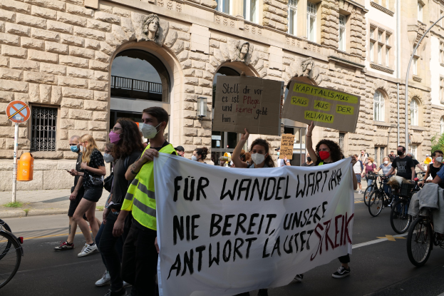 Eine Gruppe von Menschen marschiert auf einer Straße bei einer Demonstration, einige halten Schilder und andere fahren Fahrräder, im Hintergrund ein Gebäude mit Rundbogenfenstern, Säulen und Skulpturen.