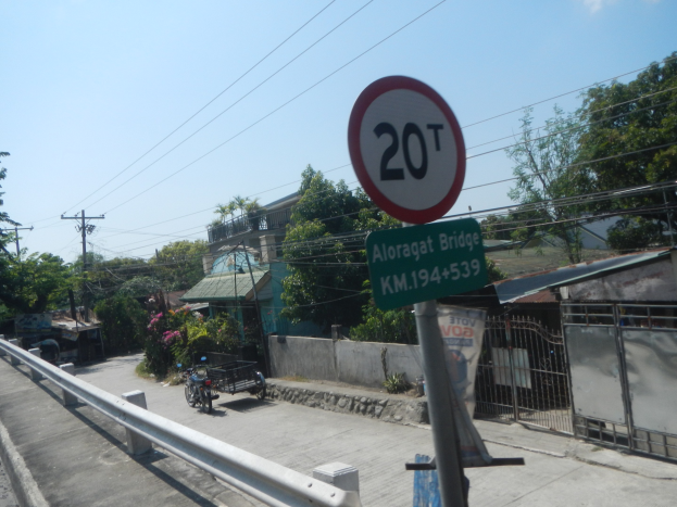Tempolimitschild am Straßenrand mit Fahrzeugen, einem Geländer, Bäumen, Gebäuden, Strommasten mit Drähten und einem bewölkten Himmel im Hintergrund.