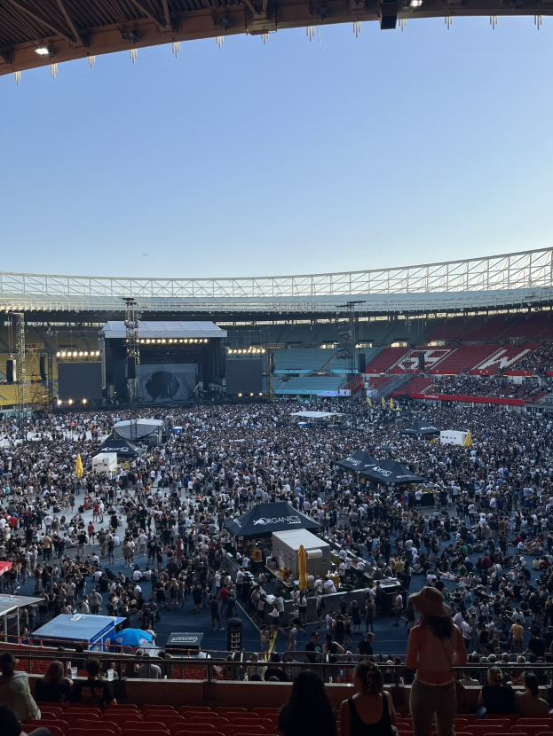 Große Menschenmenge auf Tribünen und Stehplätzen in einem Stadion mit Zelten, Geländern und Deckenbeleuchtung unter einem Dach, vor einem Himmel als Hintergrund, was auf ein Konzert in der O2 Arena in London hindeutet.