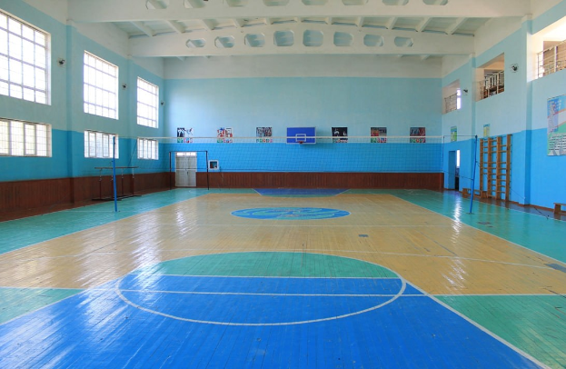 Große Turnhalle mit einem blauen und weißen Basketballfeld, einem Netz an Stangen, einer Tür, Fenstern, Wandplakaten, einem Geländer und einem Dach.